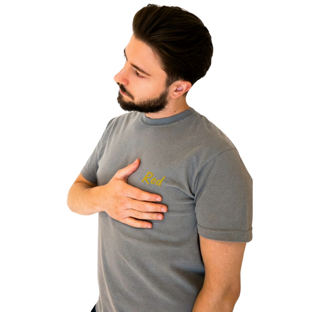 A guy wearing a Gray T-shirt with the word 'Red' embroidered in Yellow on the left chest. This soft, premium cotton T-shirt is designed for comfort and everyday style. The model poses with one hand on his chest, showcasing the embroidery and fit of the shirt with a thoughtful side profile.