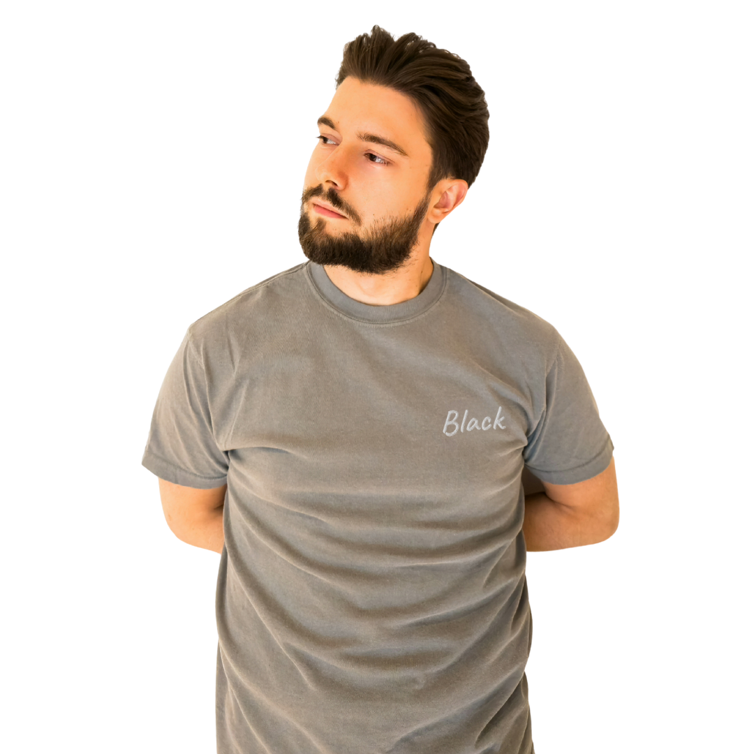 A man wearing a Gray T-shirt with the word 'Black' embroidered in White on the left chest. The T-shirt is made from premium cotton for a comfortable fit, ideal for everyday wear and casual style. The model is posing with his arms behind his back, looking to the side to highlight the shirt's front design.