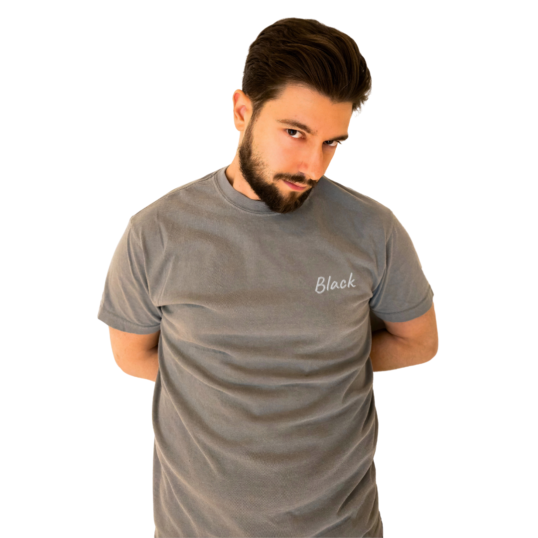 A man wearing a Gray T-shirt with the word 'Black' embroidered in White on the left chest. This high-quality cotton T-shirt is designed for a relaxed and modern fit, ideal for everyday wear. The model stands with his arms behind his back and a playful, confident look, emphasizing the shirt's unique design.