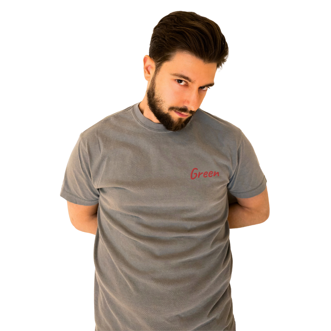 A man wearing a Gray T-shirt with the word 'Green' embroidered in Red on the left chest. This high-quality cotton T-shirt is designed for a relaxed and modern fit, ideal for everyday wear. The model stands with his arms behind his back and a playful, confident look, emphasizing the shirt's unique design.