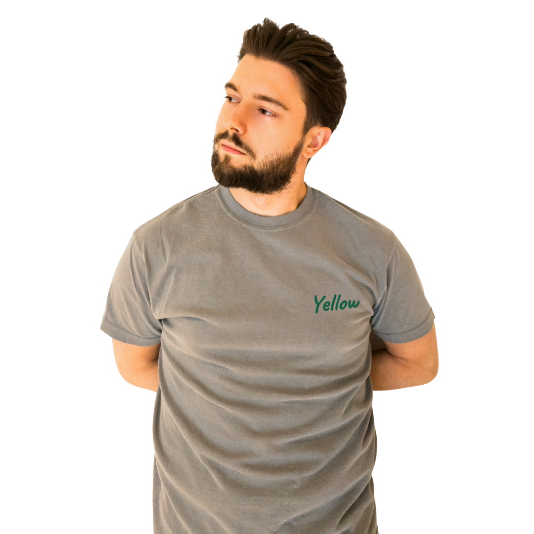 A man wearing a gray T-shirt with the word 'Yellow' embroidered in green on the left chest. The T-shirt is made from premium cotton for a comfortable fit, ideal for everyday wear and casual style. The model is posing with his arms behind his back, looking to the side to highlight the shirt's front design.