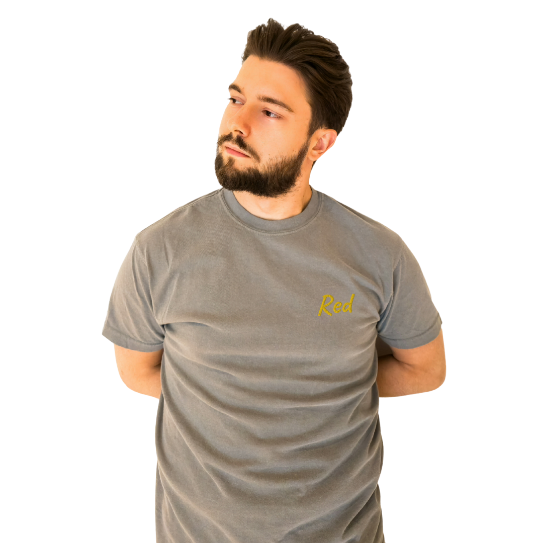 A man wearing a Gray T-shirt with the word 'Red' embroidered in Yellow on the left chest. The T-shirt is made from premium cotton for a comfortable fit, ideal for everyday wear and casual style. The model is posing with his arms behind his back, looking to the side to highlight the shirt's front design.